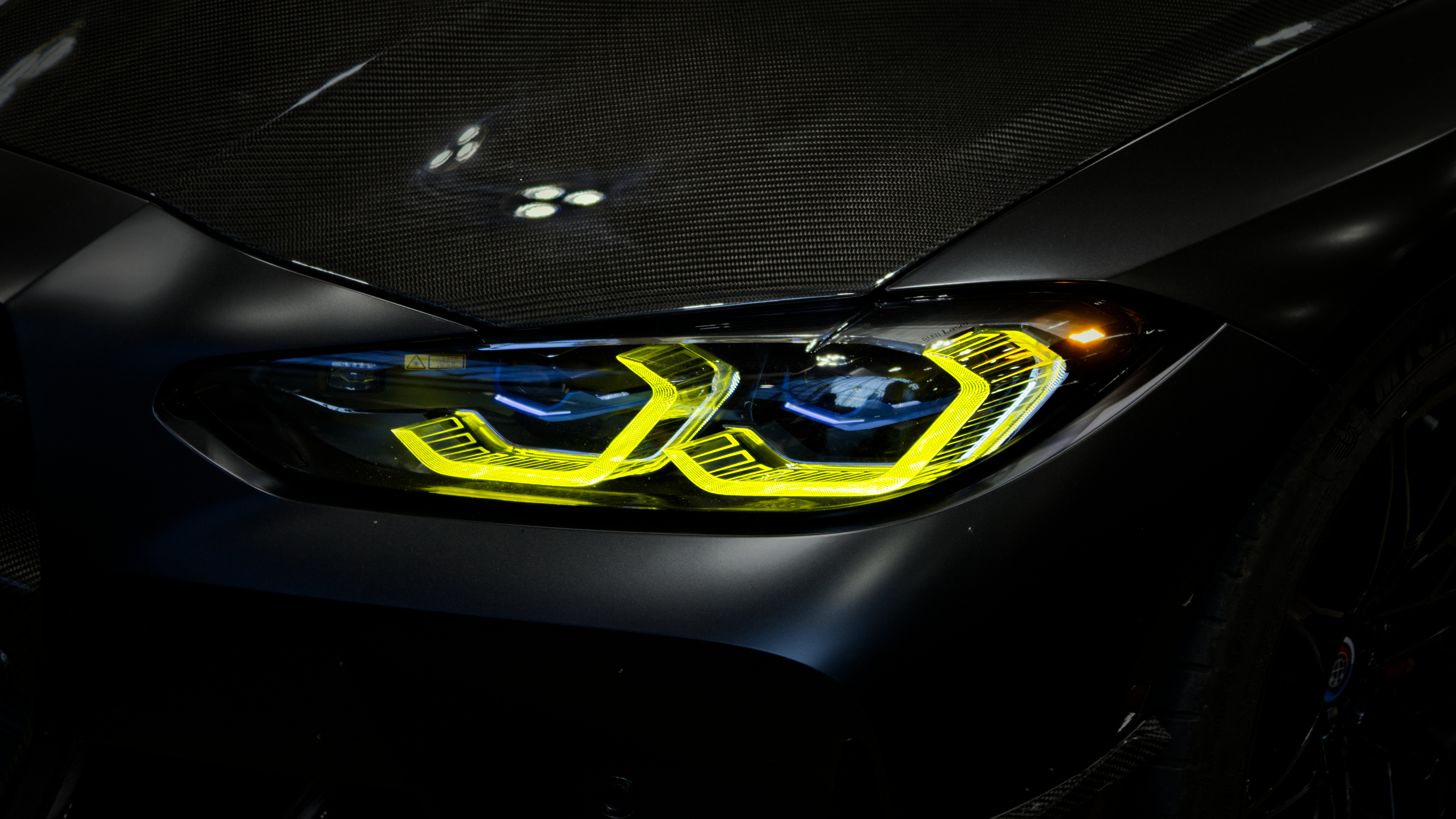 Close-up of a BMW M4 headlight with distinctive yellow laser accents on a carbon fiber hood